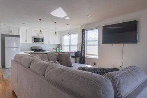 Eine moderne Küche mit weißen Schränken, einer Mikrowelle und einem Kühlschrank. Ein graues Sofa mit Kissen und ein flachbildfernseher an der Wand.