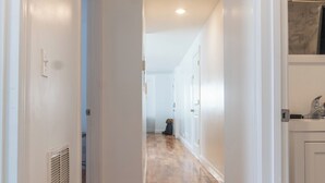 Un pasillo con piso de madera, paredes blancas y una puerta que da a una habitación con techo blanco y un respiradero.