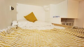 A bed with a yellow and white striped blanket, a white pillow, and a yellow patterned pillow. There is a white bedside table with a lamp and a small shelf with items on it. - Cozy Mini-Studio for 2 Near Louvre/ Metro/Elevator (Paris)