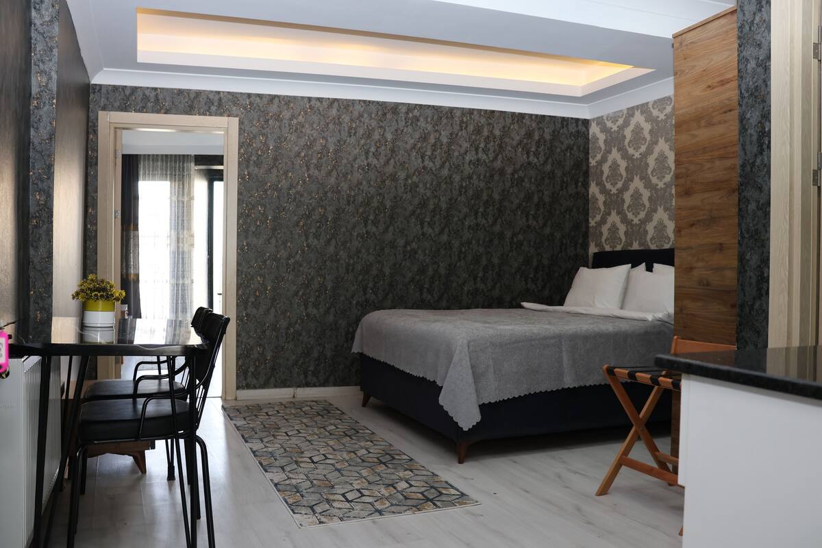 A bedroom with a bed, a desk, and a chair. There is a window with curtains, a radiator, and a small table with a plant.