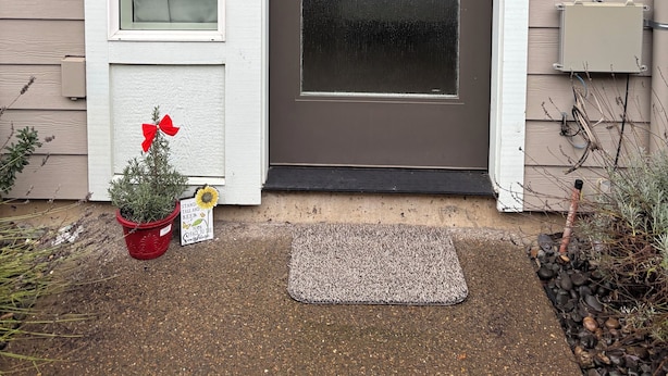 Eine Haustür mit einer Milchglasscheibe, ein kleiner Podeststufenaufgang, ein roter Blumentopf mit einer Pflanze und eine Fußmatte am Boden.