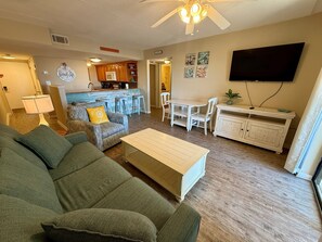 A living room with a green sofa, a coffee table, a TV, and a kitchen area with a bar stool and a microwave. - Sugar Beach 2 Bedroom, 2 Bathroom Condo. Heated Pool. Free Daily Activities! (Orange Beach)