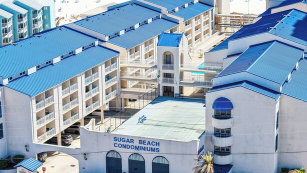 A multi-story building with a blue roof, labeled "SUGAR BEACH CONDOMINIUMS," located near a beach. - Sugar Beach 2 Bedroom, 2 Bathroom Condo.  Heated Pool.  Free Daily Activities! (Orange Beach)