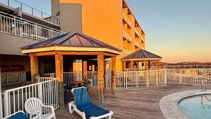 A deck with lounge chairs and a pool, surrounded by a fence and a building with balconies. - Sugar Beach 2 Bedroom, 2 Bathroom Condo.  Heated Pool.  Free Daily Activities! (Orange Beach)