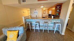 A cozy living room with a sofa and a coffee table, a kitchen with wooden cabinets and a bar area with stools. - Sugar Beach 2 Bedroom, 2 Bathroom Condo.  Heated Pool.  Free Daily Activities! (Orange Beach)