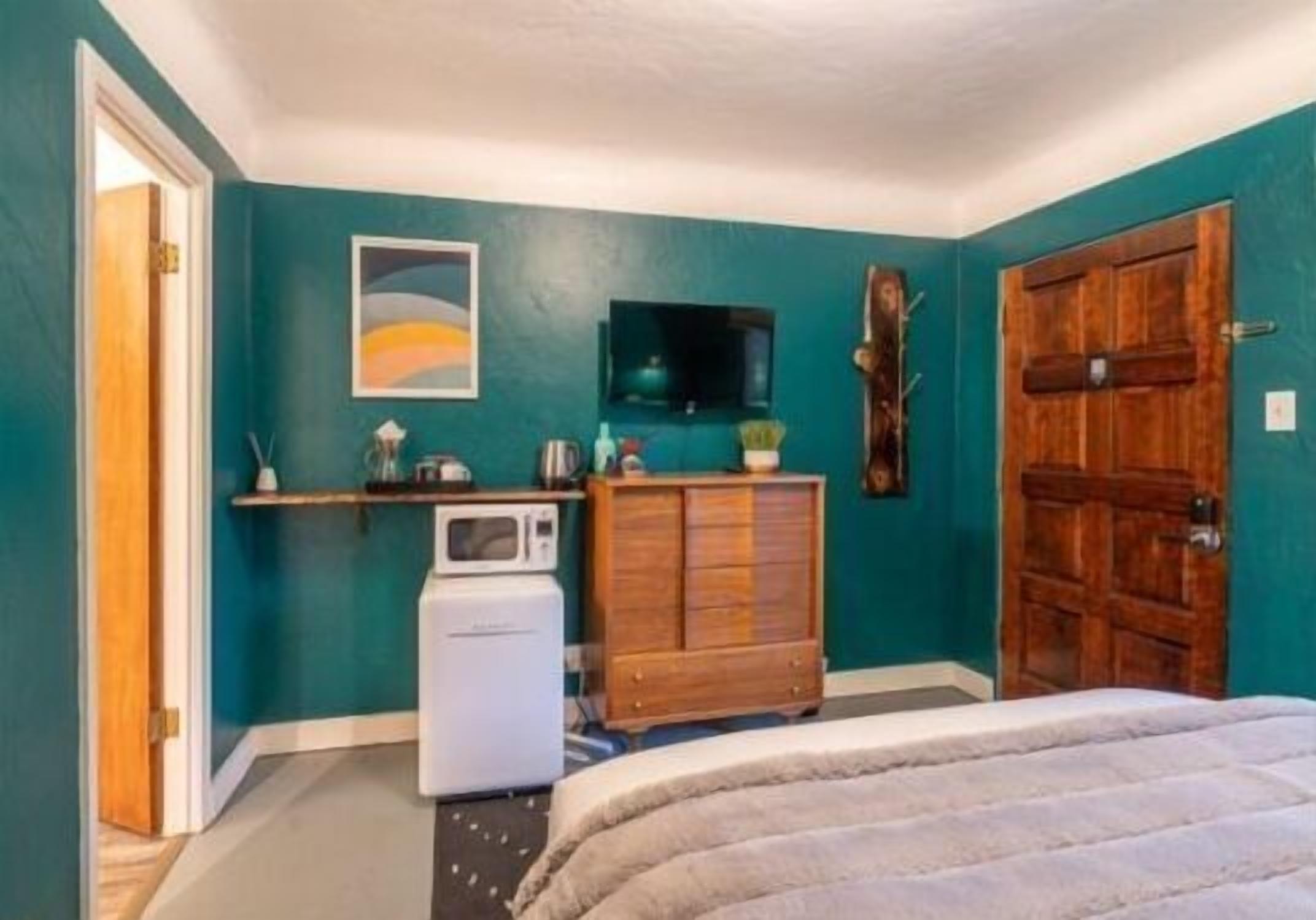 A bedroom with teal walls, a wooden door, a bed, a wooden dresser, a microwave, and a TV on the wall.