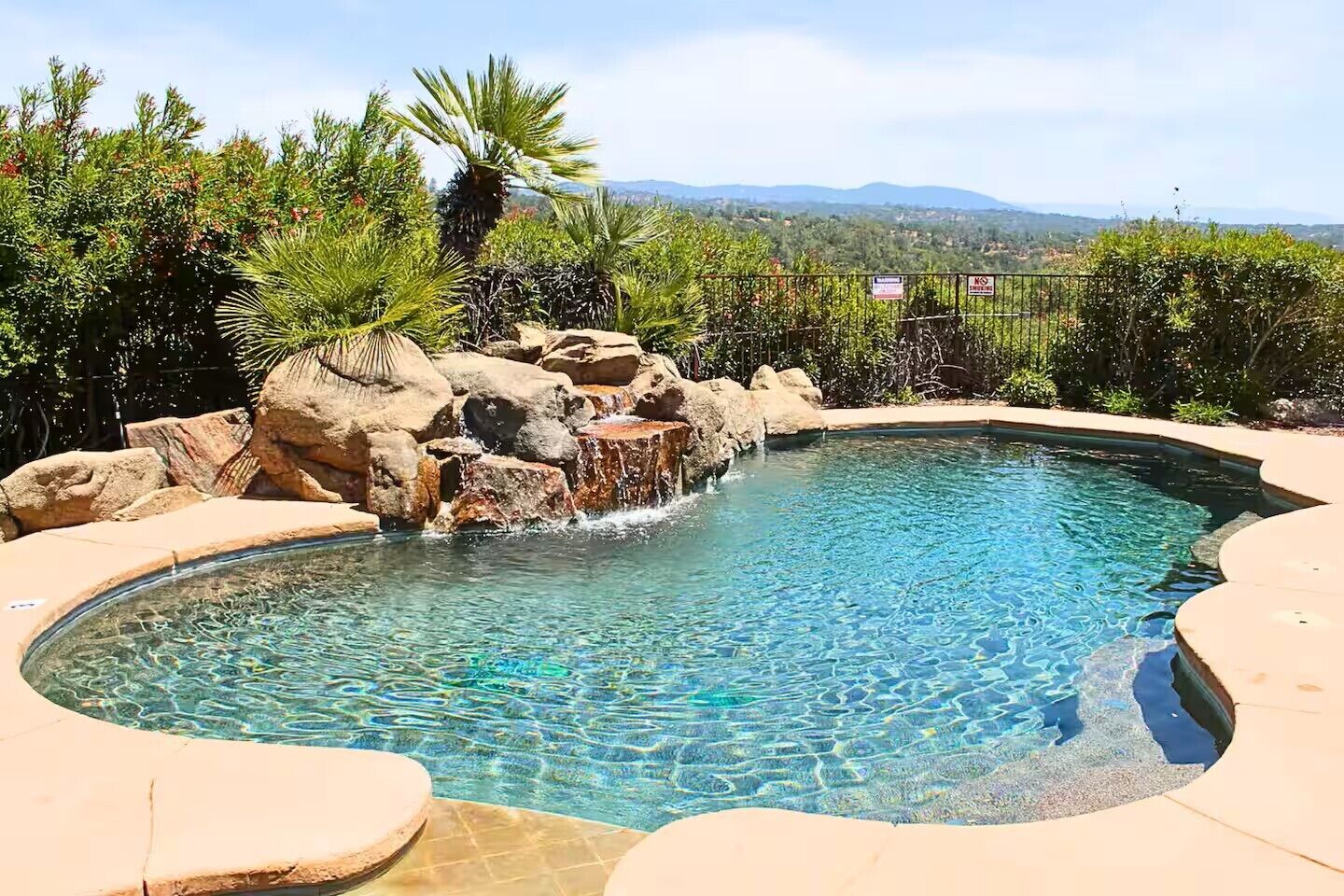 A clear swimming pool with a rocky waterfall feature, surrounded by a stone deck and lush greenery.