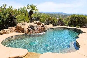 Una piscina cristalina con una cascada artificial de rocas, rodeada por una terraza de piedra y abundante vegetación.