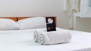 Een netjes opgemaakt bed met witte lakens en een opgevouwen handdoek, een bordje met de hotelnaam en een badjas hangend aan de muur.
