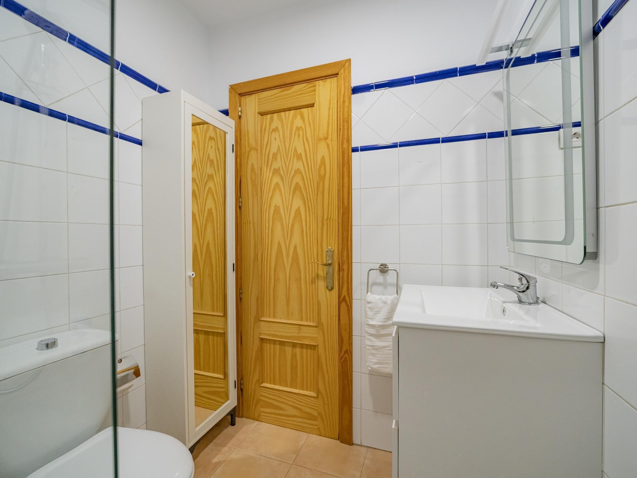 Un baño con puerta de madera, azulejos blancos, cabina de ducha con mampara de vidrio y lavabo blanco.