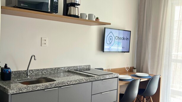 A modern kitchen with a granite countertop, a coffee maker, and a television displaying a check-in message. - Hype Vila Mariana (São Paulo)