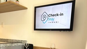A dining area with a television displaying a check-in screen, two chairs, a table with plates and a glass, and a kitchen counter in the background. - Hype Vila Mariana (São Paulo)