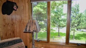 A room with a wooden interior, a window with a view of trees, a lamp, and a bear head decoration. - Nitschke's Resort Cabin #15 (Minocqua)