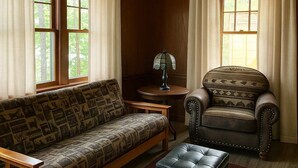 Un salon chaleureux avec un canapé à motifs, un fauteuil en cuir et un petit pouf. On y trouve une lampe sur une table d’appoint et une fenêtre avec des rideaux.