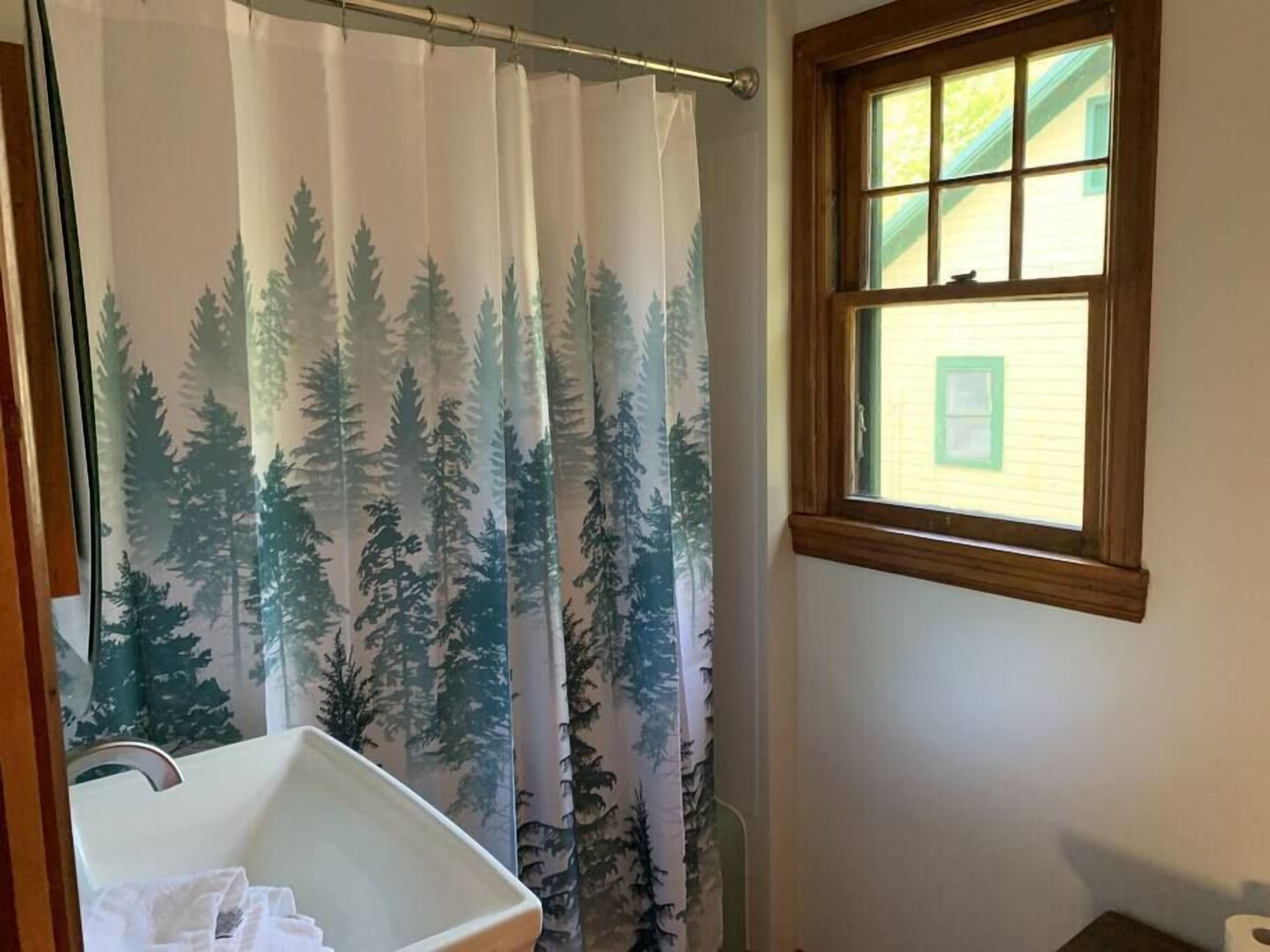 A bathroom with a shower curtain featuring a forest print, a window with a view of a house, and a white bathtub.
