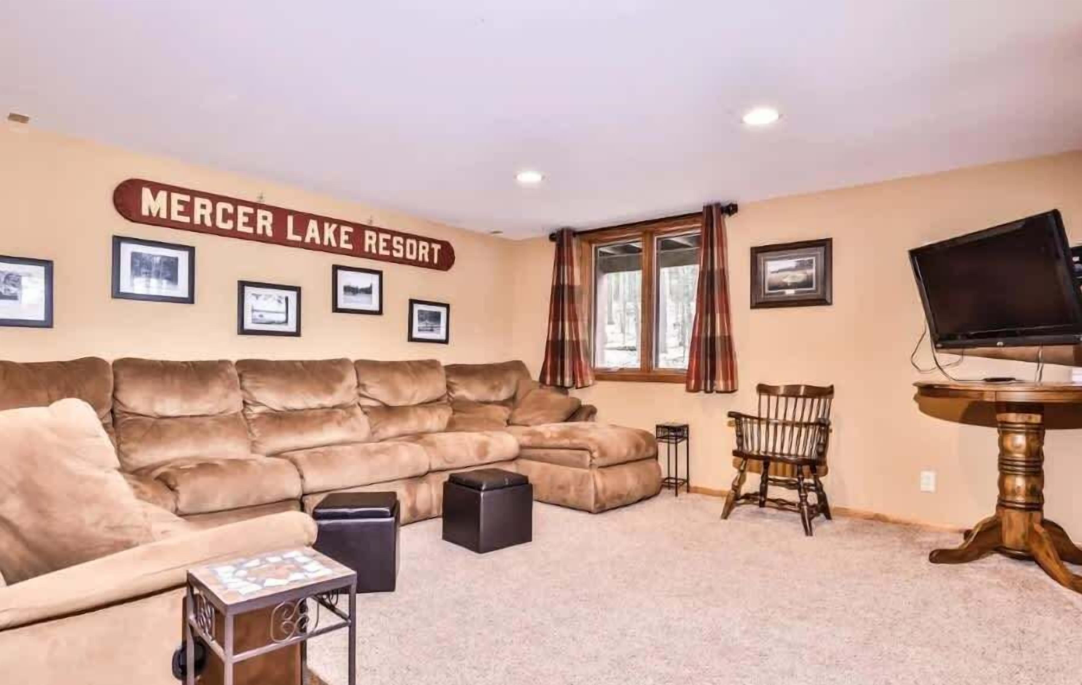 A living room with a large beige sectional sofa, a small wooden table, a television on a wooden stand, and a sign reading "Mercer Lake Resort" on the wall.