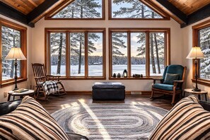 A cozy room with large windows, a rocking chair, a blue armchair, and a circular rug. - Mercer Lake Musky Lodge (Minocqua)