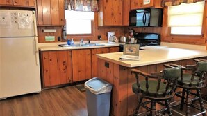 A wooden cabin-style kitchen with a refrigerator, stove, microwave, and a small island with a laptop. There are wooden chairs and a trash can. - Old Stoney's Bass (Woodruff)