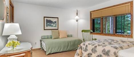 Ein Schlafzimmer mit zwei Betten, einem Schreibtisch mit Lampe, einem gerahmten Bild an der Wand und einem Fenster mit Blick auf Bäume.