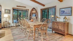 Spiseområde med et træbord og stole, en rustik kommode og et stort vindue med udsigt til træer.