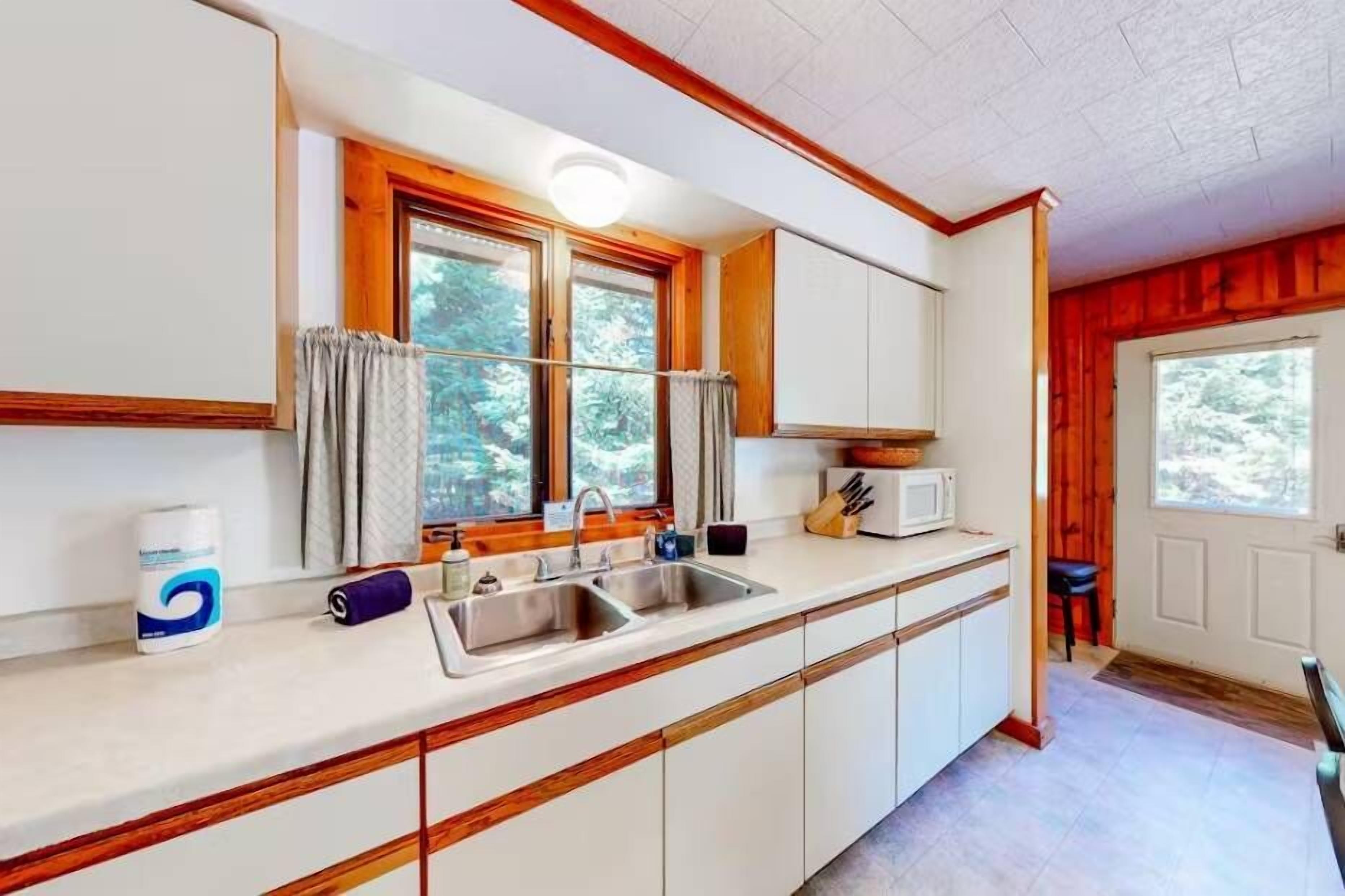 A kitchen with white cabinets, a double sink, a microwave, and a window with curtains.