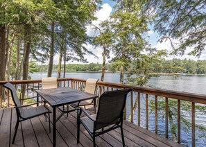 Een houten terras met een tafel en stoelen met uitzicht op een meer, omringd door hoge bomen.