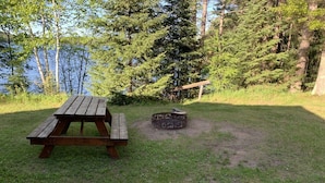 A picnic table and a fire pit in a grassy area with trees and a body of water in the background. - Gresham Shores - Smiling Moose - PET Friendly (Woodruff)