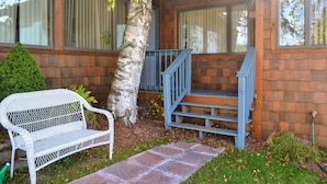 Une maison en bois avec un escalier bleu menant à un porche. On y trouve un banc en osier blanc et un petit arbre devant.