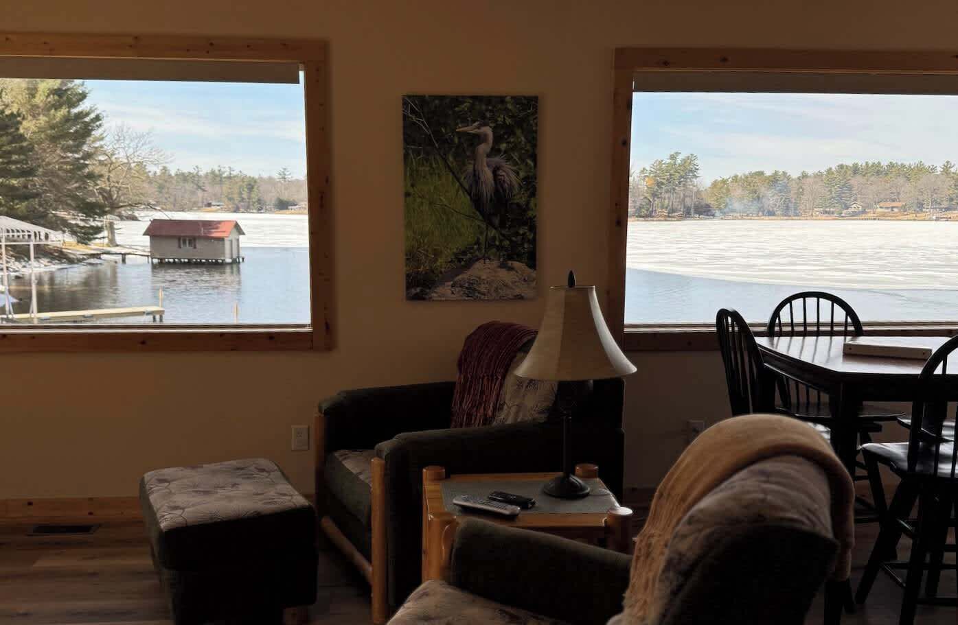 A cozy living room with a view of a lake, a painting on the wall, and a dining area with chairs and a table.
