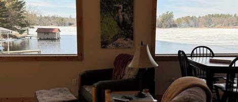 A cozy living room with a view of a lake, a painting on the wall, and a dining area with chairs and a table.