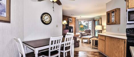 A dining area with a wooden table and white chairs, a ceiling fan, a clock, and a kitchen with wooden cabinets and a stove.