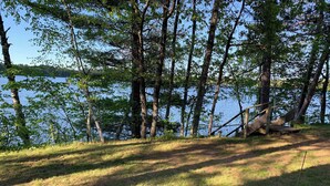 Un paysage naturel avec un sentier de terre menant à une structure en bois située près d’une étendue d’eau, le tout entouré d’arbres.