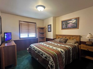 A bedroom with a bed, a desk, a computer, a window with blinds, a shelf, a lamp, and a framed picture on the wall. - Northern Resort #17 (Minocqua)