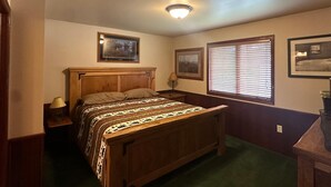 Ein Schlafzimmer mit einem Holzbett, einem Nachttisch, einer Lampe, einem gerahmten Bild an der Wand und einem Fenster mit Jalousien.