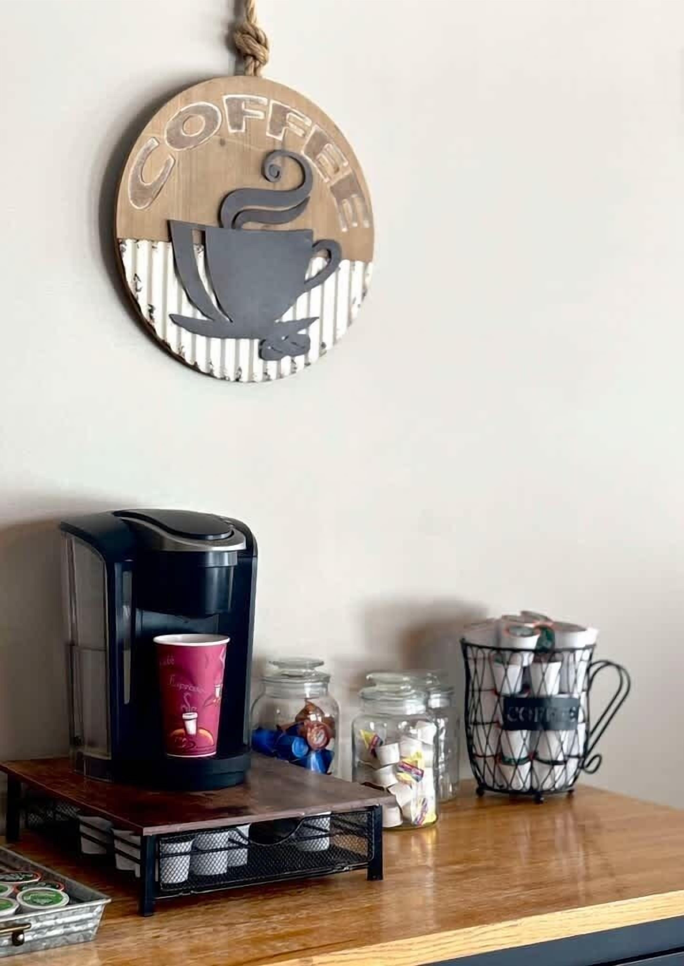 A coffee maker on a wooden table with a coffee cup, jars of coffee beans, and a coffee-themed wall hanging.