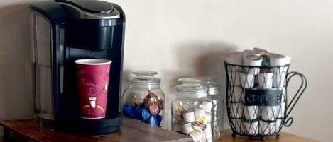 A coffee maker on a wooden table with a coffee cup, jars of coffee beans, and a coffee-themed wall hanging.