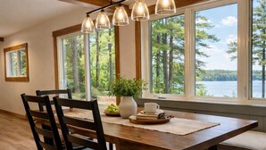 A dining area with a wooden table, chairs, and a view of a lake through large windows. - Moss Lake Lookout: PET Friendly (Lac du Flambeau)