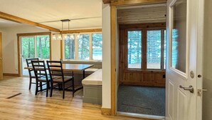 A spacious room with a wooden dining table and chairs, large windows with a forest view, and a door leading to another area. - Moss Lake Lookout: PET Friendly (Lac du Flambeau)