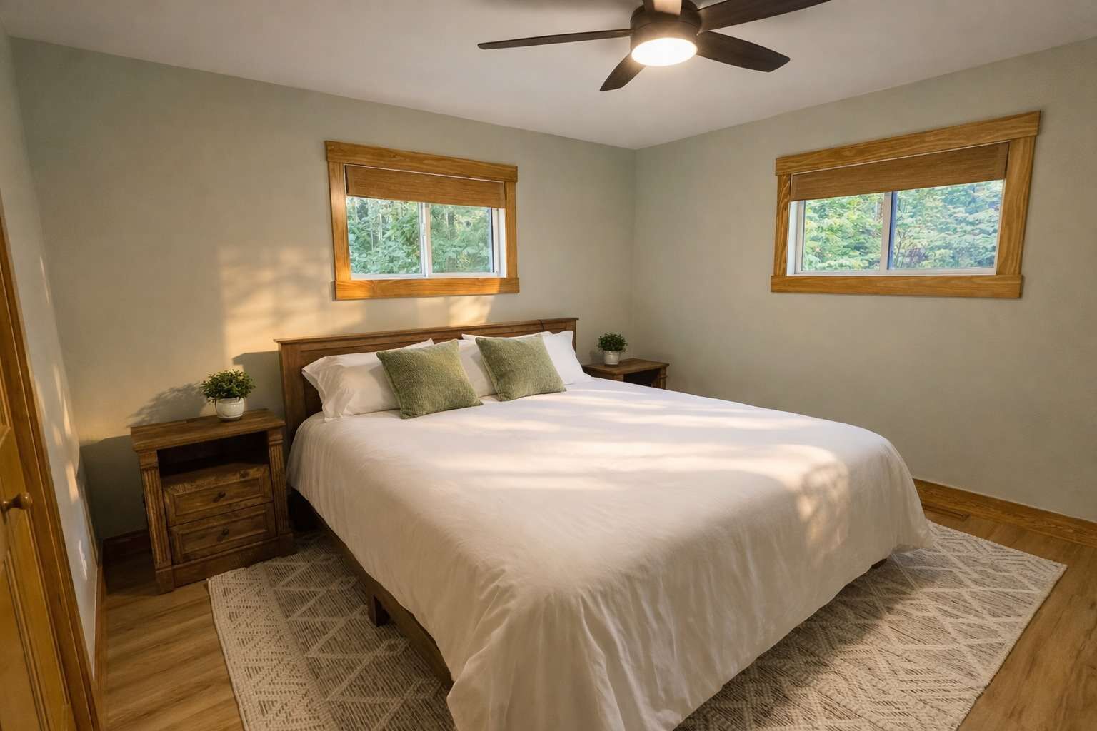 A bedroom with a bed, wooden headboard, two nightstands, a ceiling fan, and two windows with wooden frames.