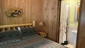 Una habitación con paneles de madera, con una cama que tiene ropa de cama a cuadros y estampados de animales, un cabecero de madera, una mesita de noche con una lámpara y una puerta que da a un baño.