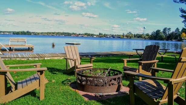 A lakeside deck with wooden chairs and a fire pit, overlooking a calm lake with a dock and picnic tables. - Minocqua Shores Cabin #2 (Minocqua)