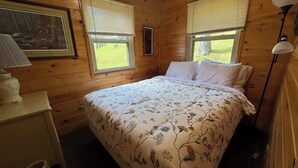 A wooden cabin bedroom with a bed, a nightstand, a lamp, and a window with blinds. - Minocqua Shores Cabin #2 (Minocqua)