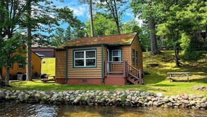 A log cabin with a porch and a small staircase, situated next to a body of water and surrounded by trees. - Minocqua Shores Cabin #2 (Minocqua)