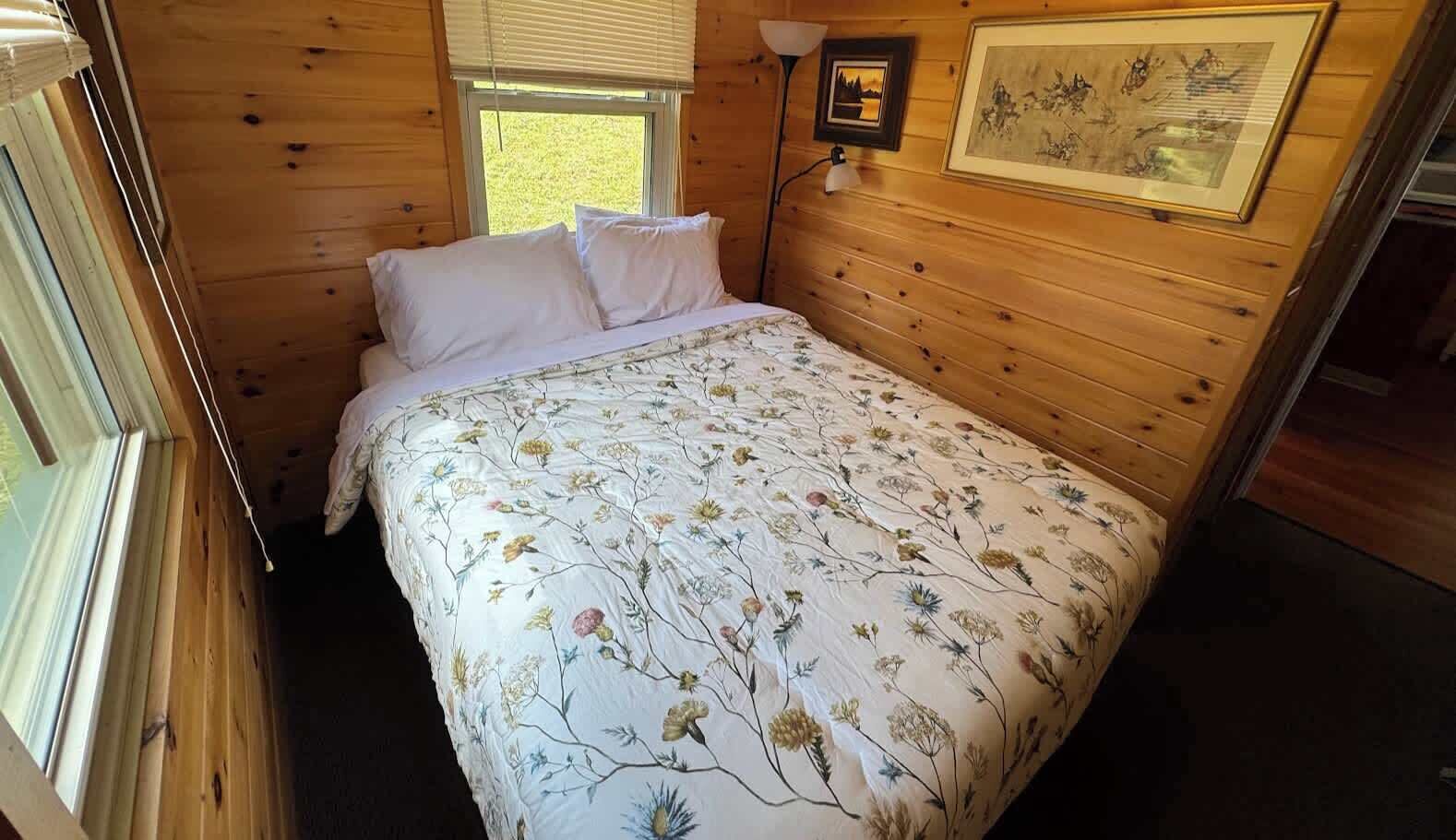A wooden cabin bedroom with a bed, two pillows, a window, and a wall-mounted lamp.