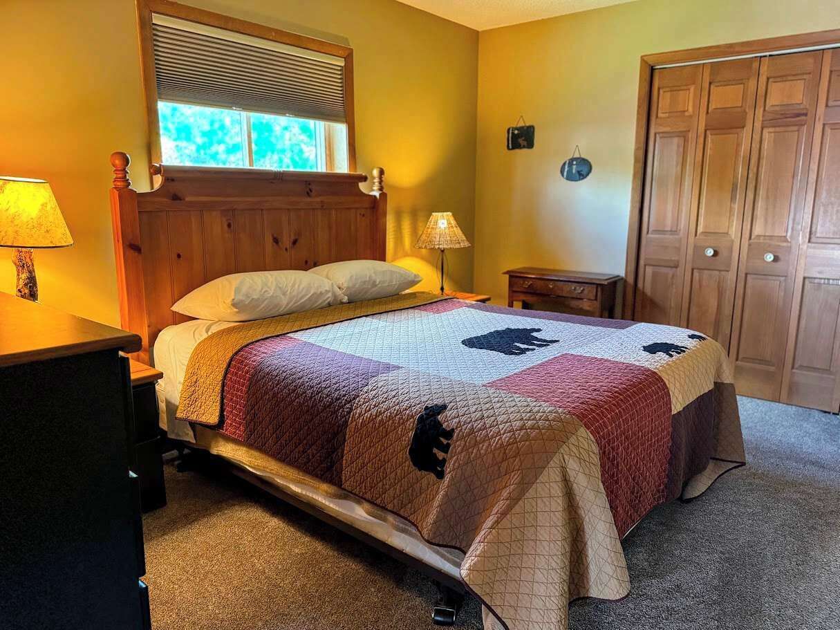 Une chambre à coucher comprenant un lit, une tête de lit en bois, des tables de chevet, une fenêtre avec des stores, une armoire en bois et une horloge murale.