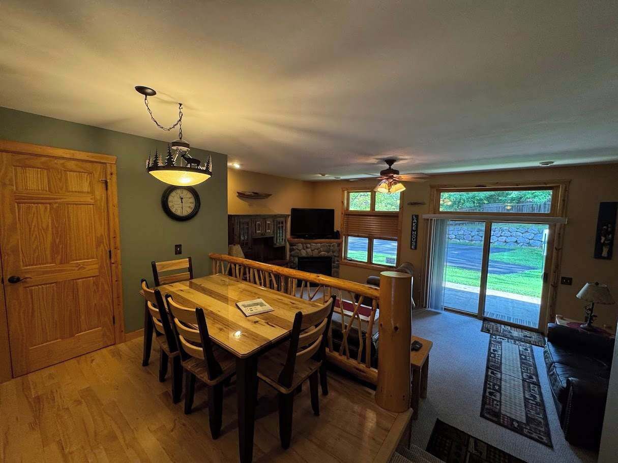 A dining area with a wooden table and chairs, a clock on the wall, and a television in the background.