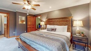 Un dormitorio con una cama grande, cabecera de madera, dos lámparas de noche, un ventilador de techo y un banco de madera al pie de la cama.