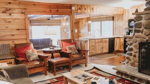 A cozy cabin interior with wooden walls, a stone fireplace, a dining area with a table and chairs, and a kitchen with wooden cabinets and a stone countertop. - Decompression Zone: Direct Access TO Snowmobile Trails (Woodruff)