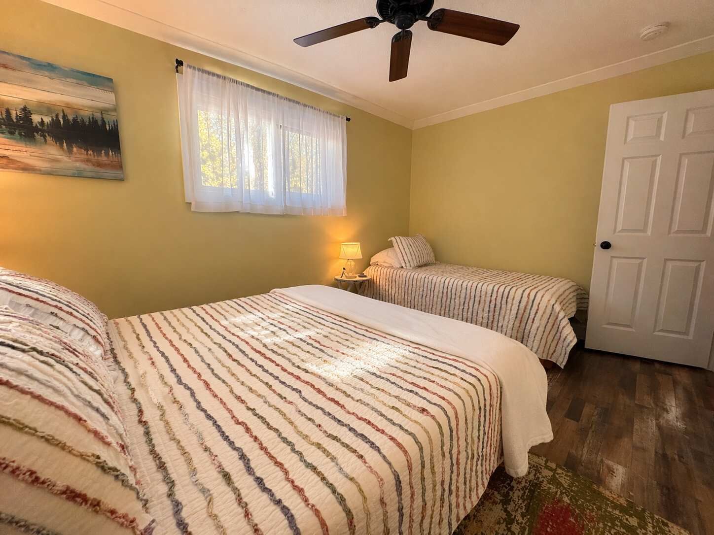 Una habitación con dos camas, un ventilador de techo, una ventana con cortinas traslúcidas y una puerta.