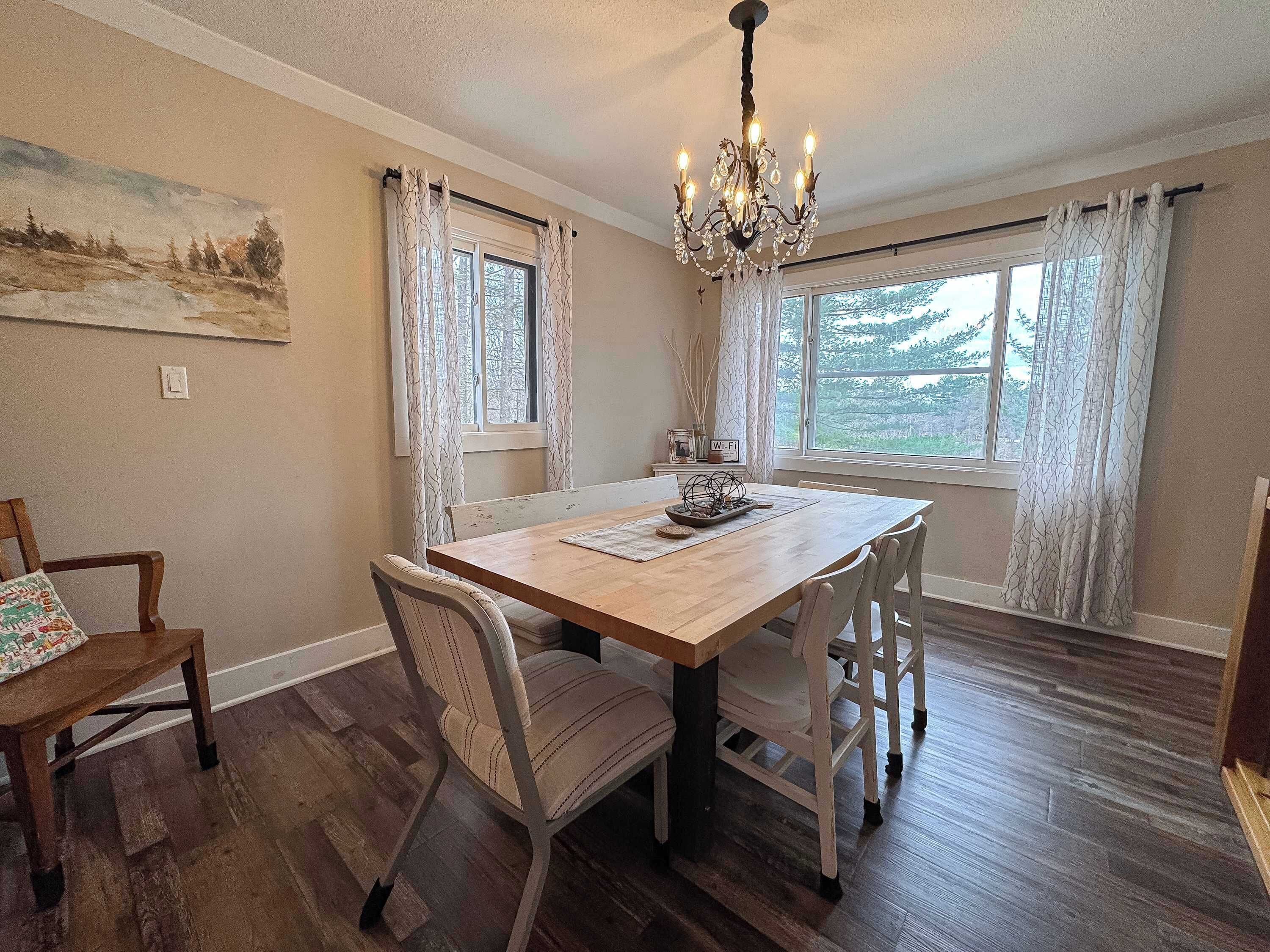 A dining room with a wooden table, chairs, a chandelier, and large windows with curtains.